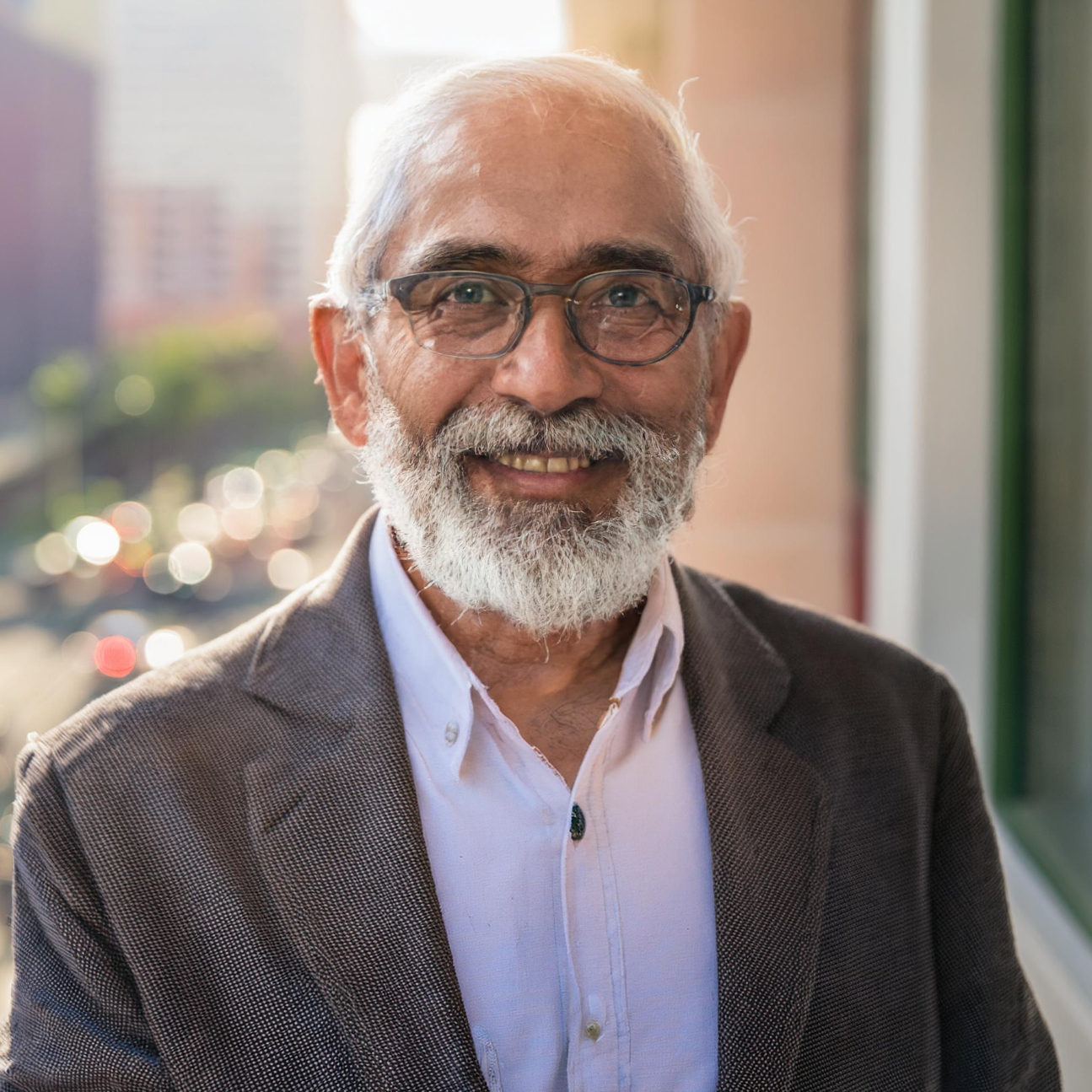Photo of a smiling man wearing a suit and with glasses, stood in front of a window 